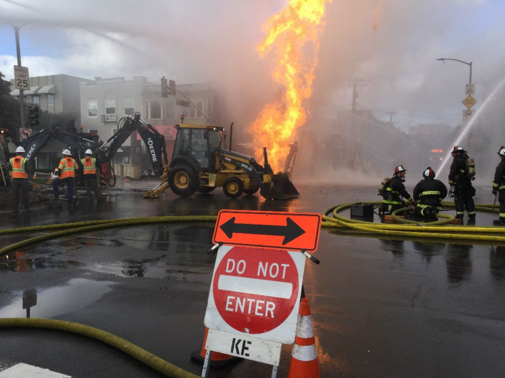 An outdoor fire burns in front of a mini excavator amid smoke as firefighters try to extinguish the flames.